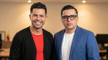 Dos hombres, Piter Albeiro sonriente y Beto Coral con gafas, posan en una oficina. Albeiro viste cárdigan negro y camiseta roja; Coral, blazer azul y camiseta blanca.