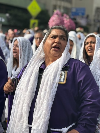 Una mujer en primer plano y otras detrás visten túnicas moradas y mantillas blancas de encaje mientras cantan en una calle concurrida