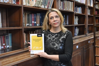 Mujer con cabello rubio, jersey negro con encaje, pendientes dorados, sentada ante estanterías de madera llenas de libros, con un libro amarillo