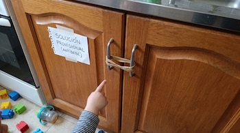 Puertas de madera de un mueble de cocina sujetas por una banda elástica en los tiradores, un cartel pegado a la izquierda y la mano de un niño.