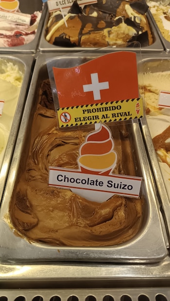Bandeja metálica con gelato de chocolate. Un cartel muestra la bandera de Suiza y el texto 'Chocolate Suizo' y 'PROHIBIDO ELEGIR AL RIVAL'