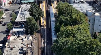 Vista aérea de un tren en vías de una estación con plataformas amarillas, árboles, edificios con techos planos, una calle con vehículos y personas