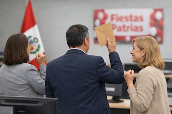 Un hombre y dos mujeres en una oficina. El hombre sostiene un sobre. Se ve una bandera de Perú y un cartel de Fiestas Patrias de fondo, con monitores.