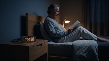 Un reloj digital sobre una mesa de noche marca las 03:00 junto a un hombre en pijama sentado en la cama leyendo un libro