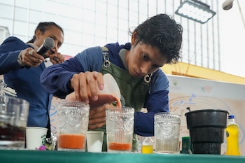 Un hombre con delantal azul y verde vierte un líquido de color rojizo en vasos de cristal sobre una mesa. Otro hombre sostiene un micrófono en el fondo