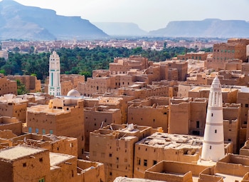 Vista aérea de una ciudad de adobe con dos minaretes blancos. Al fondo se observa un oasis con palmeras y varias cadenas montañosas