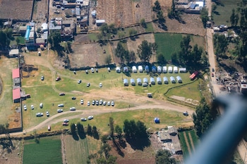 Vista aérea de un terreno verde con hileras de carpas blancas, automóviles estacionados y campos agrícolas