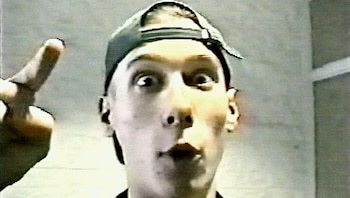 Primer plano de un joven con gorra al revés, ojos abiertos y boca sorprendida, apuntando hacia arriba con su dedo índice izquierdo. La imagen tiene baja calidad de video