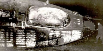 Fuselaje de avión Tupolev Tu-134 dañado con ventanillas rotas, una persona de pie junto al fuselaje y fondo oscuro. Imagen en blanco y negro