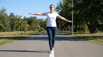 Mujer con camiseta blanca y jeans oscuros caminando sobre una línea blanca en un sendero de asfalto con brazos extendidos; fondo de parque con árboles y césped.