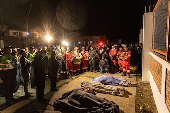 Agrupación de bomberos, policías y civiles en la noche; tres cuerpos cubiertos con mantas en el suelo; luces de emergencia y un edificio con pared de ladrillo