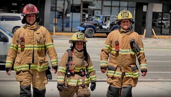 Tres bomberos con trajes de protección y cascos caminan por la calle frente a vehículos estacionados y un edificio