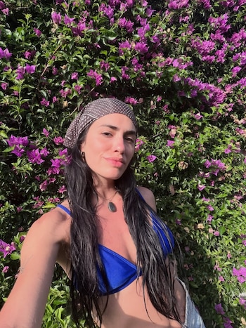 Mujer con cabello oscuro mojado, bikini azul, pañuelo de puntos y collar con una piedra, posa frente a un seto de buganvilias de color púrpura