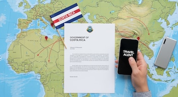 Vista cenital de un escritorio con un mapa del Medio Oriente, una carta oficial del Gobierno de Costa Rica, una bandera costarricense y una mano con un teléfono que muestra "TRAVEL ALERT".