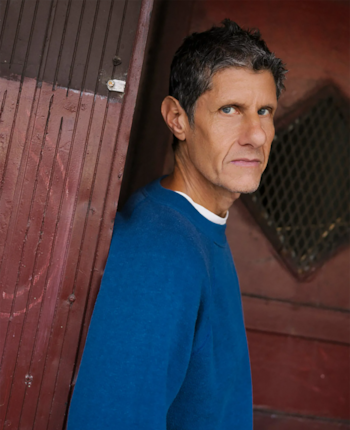 Retrato de un hombre de cabello gris y ojos azules, con sudadera azul y camiseta blanca, apoyado en una puerta de madera rojiza con una rejilla