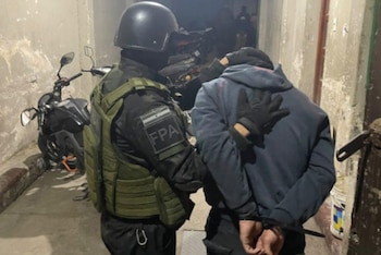 Un agente uniformado con casco y chaleco táctico sujeta a un hombre esposado que viste un buzo azul con capucha. Al fondo hay una motocicleta