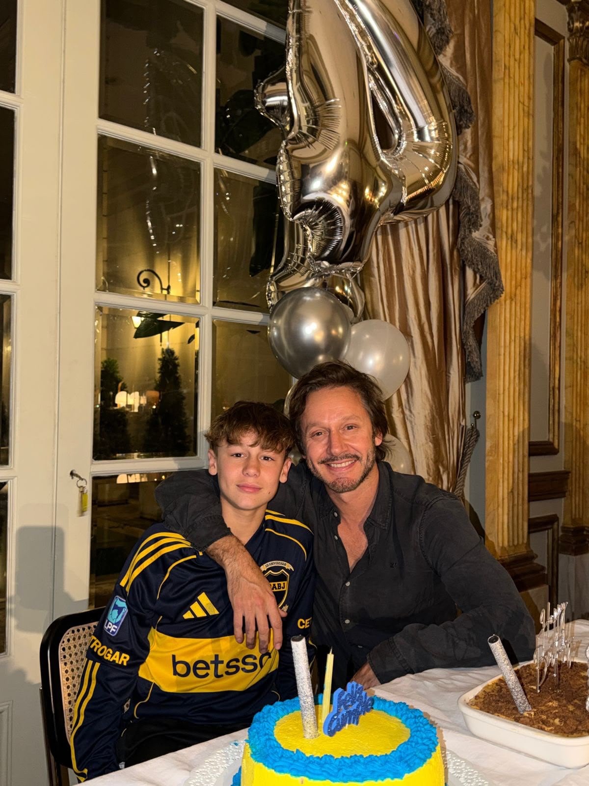 Benjamín Vicuña posa sonriente junto a su hijo, que luce una camiseta de fútbol, frente a una torta de cumpleaños y globos festivos