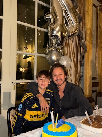 Un hombre adulto y un joven sonríen frente a una torta de cumpleaños con velas, globos plateados y un postre sobre una mesa, con una ventana de fondo