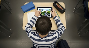 Vista cenital de un adolescente de espaldas con camiseta a rayas sentado en un pupitre, usando una tableta. Hay una cartuchera, libros y papeles en la mesa.