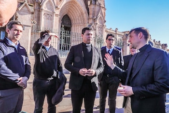 Varios hombres con trajes oscuros y alzacuellos eclesiásticos hablan frente a la fachada y las puertas de una catedral de estilo gótico