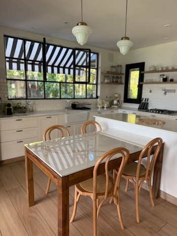 Vista interior de una cocina moderna con una mesa de madera y sillas, gabinetes blancos, un gran ventanal con toldo a rayas y electrodomésticos