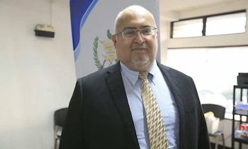 Un hombre mayor con gafas, traje oscuro, camisa azul y corbata posa de pie frente a un cartel institucional