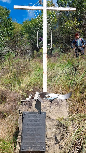 Cruz de metal blanca, placa conmemorativa, restos metálicos, base de piedra, hierba, árboles, cielo azul, persona con mochila y gorra roja en segundo plano