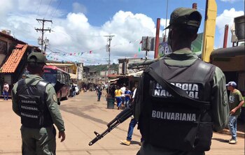 Dos miembros de la Guardia Nacional Bolivariana, de espaldas, con uniformes militares y chalecos, uno con un rifle, patrullan una calle con edificios y personas