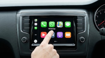 Una mano toca el icono de WhatsApp en la pantalla táctil de un sistema de infoentretenimiento de un vehículo, con otras aplicaciones visibles en el fondo oscuro.