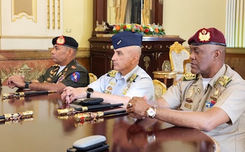Tres hombres de uniforme militar con insignias, sentados en una mesa de madera con bastones de mando, libretas y relojes de pulsera