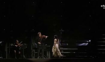 Lali Espósito, de espaldas y con vestido claro, en el escenario oscuro de River, frente a un violinista. Otro músico toca a la izquierda, con escaleras detrás