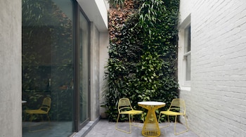 Un patio exterior moderno presenta un gran jardín vertical con follaje verde, una pared de ladrillo blanco y un juego de mesa y dos sillas de metal amarillo.
