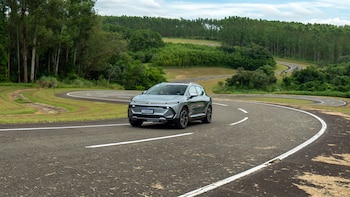 Un SUV eléctrico gris claro circula por una carretera asfaltada con curvas en un entorno verde y montañoso bajo un cielo nublado
