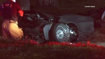 Un automóvil deportivo gris completamente destruido junto a un árbol durante la noche, con un socorrista parado al lado izquierdo