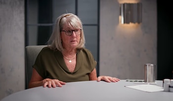 Mujer senior con cabello gris y gafas sentada en una mesa. Sobre la mesa hay una taza metálica, frascos blancos, papeles y objetos pequeños