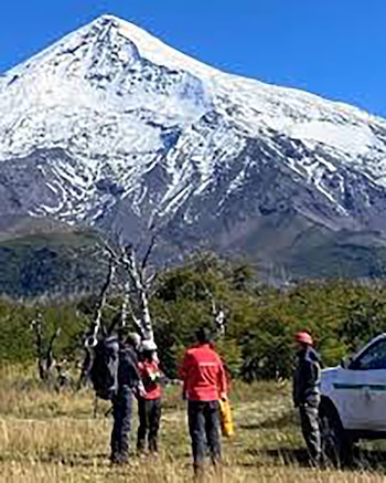 Vista panorámica del volcán Lanín cubierto de nieve bajo un cielo azul. En primer plano, varias personas con equipo de montaña y un vehículo blanco