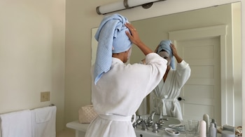 Vista posterior de una persona en albornoz blanco y toalla azul en la cabeza, aplicándose una mascarilla facial frente al espejo de un baño.