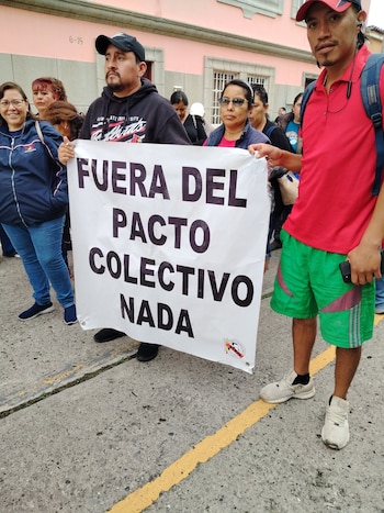 Un grupo de personas en la calle. Dos hombres sostienen una pancarta blanca con texto negro: FUERA DEL PACTO COLECTIVO NADA. Un edificio color salmón de fondo