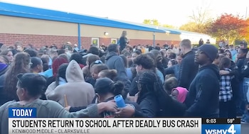 Una multitud de estudiantes y adultos se congrega frente a un edificio de ladrillo rojo y un techo azul bajo un cielo claro