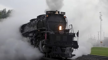 Una locomotora de vapor negra con la inscripción X4014 y una pequeña bandera estadounidense avanza sobre vías en un entorno de árboles verdes