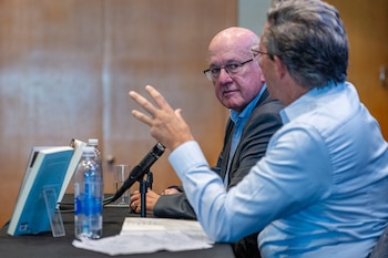 Pablo Sirvén, un hombre calvo con gafas y camisa azul, sentado a una mesa, mira a un hombre de espaldas que gesticula. Sobre la mesa hay un micrófono, un libro y botellas de agua