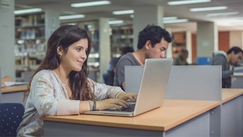 Una mujer joven con cabello largo y oscuro teclea en una computadora portátil plateada sentada en un escritorio en una biblioteca con otros estudiantes al fondo