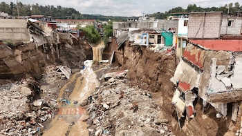Un río de agua marrón fluye entre dos laderas erosionadas donde hay casas de lámina y concreto construidas cerca del precipicio