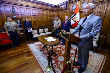 Juan Carlos Lizarzaburu habla en un podio con micrófono, junto a otros funcionarios y militares, en una sala con paneles de madera y alfombra, con la bandera peruana al fondo