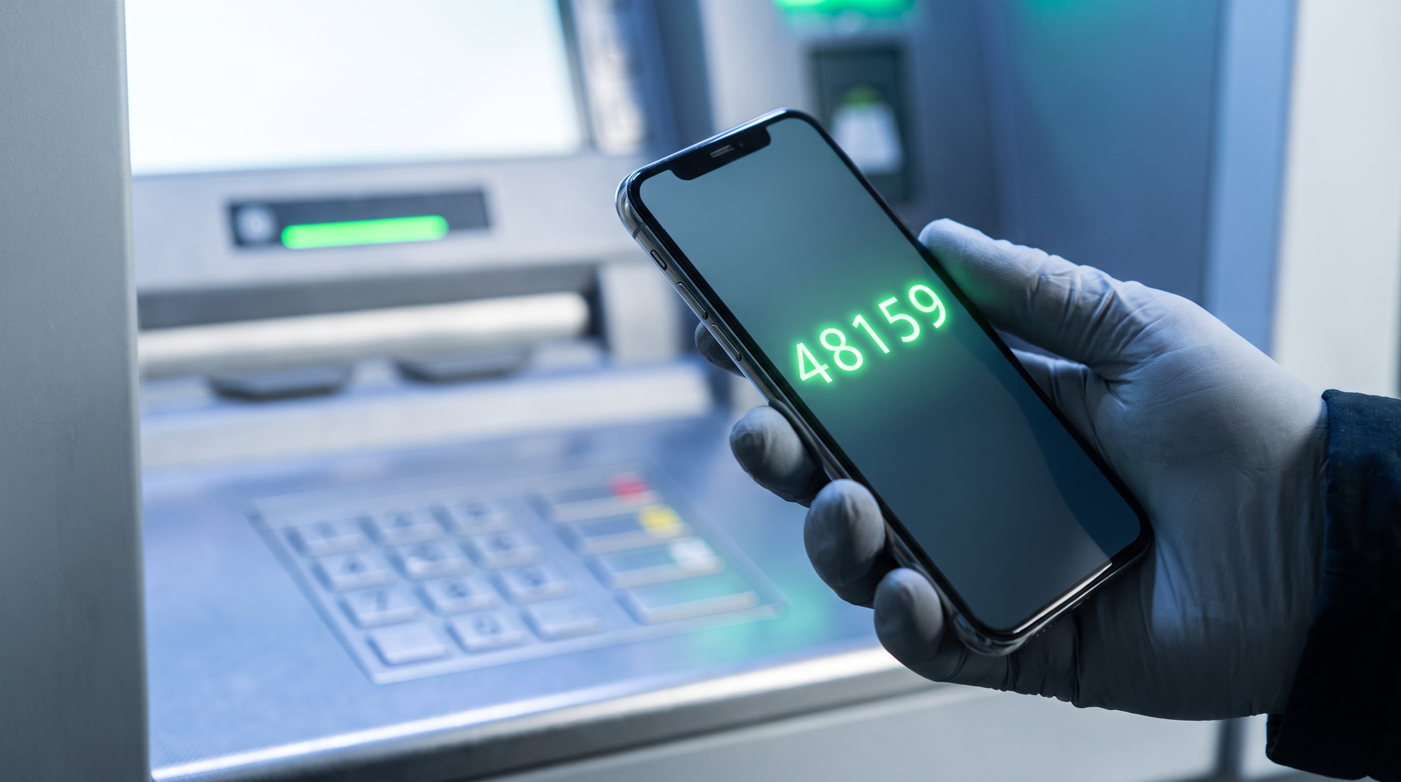 Una mano enguantada sujeta un teléfono móvil con un código numérico visible en la pantalla frente a un cajero automático. (Imagen Ilustrativa Infobae)