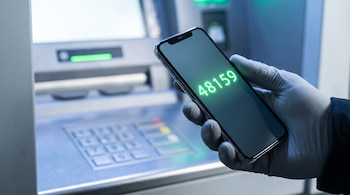 Mano enguantada sosteniendo un teléfono móvil con pantalla mostrando el número 48159 frente a un cajero automático desenfocado.