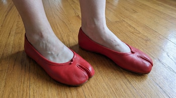 Primer plano de los pies de una persona usando zapatos planos de cuero rojo brillante con el diseño tabi, parados sobre un piso de madera.