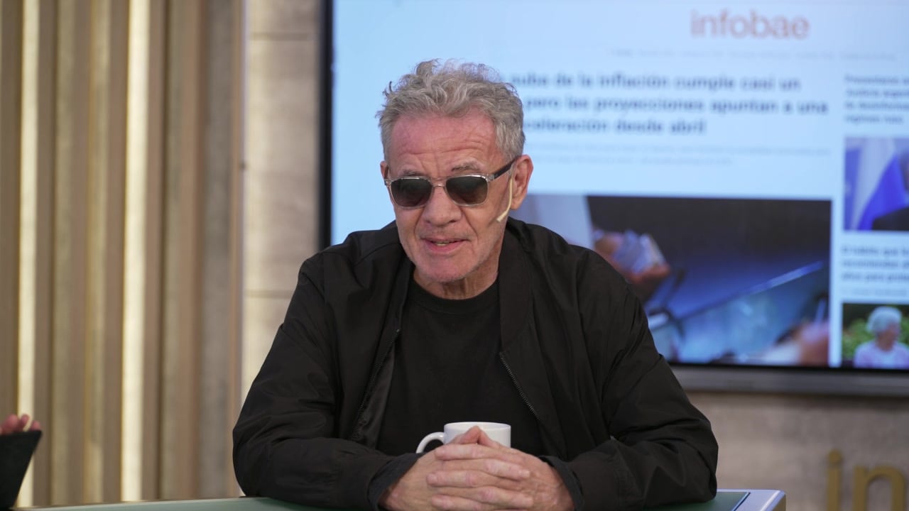 Un hombre de cabello gris y gafas de sol está sentado frente a una pantalla grande que muestra el portal de noticias Infobae, con las manos entrelazadas sobre una mesa, durante una entrevista o aparición pública.