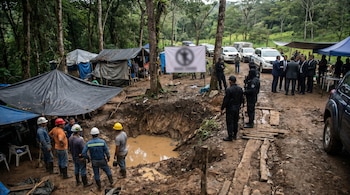 Grupo de hombres, incluyendo mineros con cascos y personal de seguridad, se congregan en un área boscosa con un pozo fangoso y campamentos improvisados.