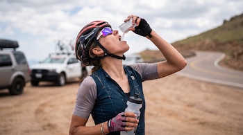 Una ciclista con casco y gafas de sol vierte polvo blanco de un sobre en su boca mientras sostiene una botella de agua.